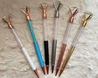 May include: A collection of six decorative pens with diamond-shaped tops. The pens have clear barrels filled with sparkling crystals and come in various colors, including white, blue, black, silver, rose gold, and gold. The pens are displayed on a white, textured surface.