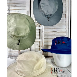 May include: Three different styles of sun hats. The first hat is a light green sun hat with a black drawstring. The second hat is a light gray sun hat with a black drawstring. The third hat is a dark blue sun hat with a red and white logo that says "It's R Thing Embroidery & Laser Design".