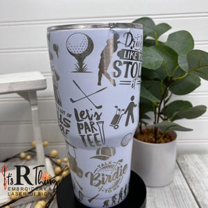 May include: White stainless steel tumbler with a golf-themed design. It features a variety of golf-related graphics and text, including "Let's Par Tee", "Drive Like You Stole It", and "Talk Birdie To Me".