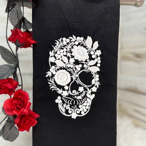 May include: Black hand towel with a white embroidered skull design. The skull is decorated with floral and leaf patterns. The towel is hanging on a silver bar, with red roses and black leaves in the background.