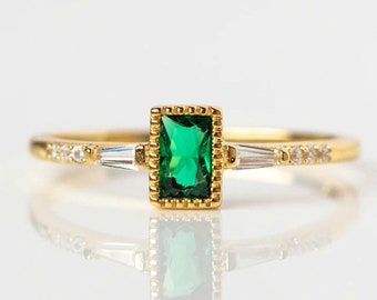 May include: A gold ring with a green emerald-cut gemstone set in a gold frame. The ring is accented with two smaller clear gemstones on either side of the emerald.