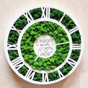 Puede incluir: Reloj redondo blanco con marcas de números romanos. La cara del reloj está cubierta de musgo verde y presenta el texto "El ingrediente secreto siempre es el amor" en español.