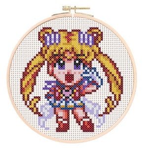 May include: Cross-stitch embroidery hoop featuring a pixelated image of Sailor Moon in her iconic pose, with her signature blonde hair, blue and red outfit, and a pink heart on her chest.