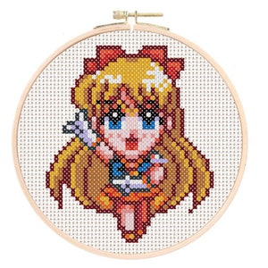 May include: A cross-stitch embroidery of a cartoon character with blonde hair, blue eyes, and a red bow. The character is wearing a blue and orange outfit, held within a wooden embroidery hoop. The background is a light beige fabric.