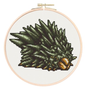 May include: A cross-stitch embroidery of a spiky, dark green creature with a golden-brown head, set in a wooden embroidery hoop. The creature's body is detailed with various shades of green, creating a textured appearance against the white fabric.