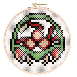 Op de afbeelding: Een borduurring met kruissteek met een gepixeld ontwerp van een groen, rood en beige wezen. Het ontwerp staat tegen een witte achtergrond en wordt omlijst door een houten ring. De kruissteek is een handgemaakt ambacht.