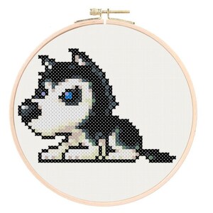 May include: Cross-stitch embroidery hoop featuring a cartoon husky dog with black and white fur and blue eyes. The dog is lying down with its head turned to the side.