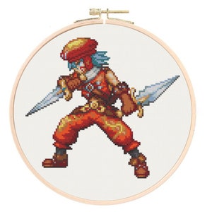May include: Cross-stitch embroidery of a pixelated warrior figure in a wooden hoop. The warrior is clad in red and brown armour, holding two silver daggers. The figure has blue hair and a red helmet. The background is white.