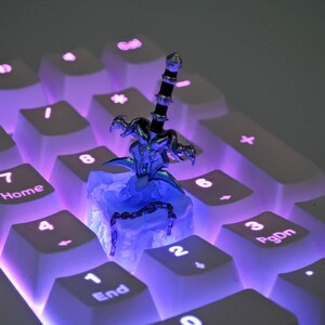 May include: A silver and black sword figurine with a blue and white base, sitting on a white keyboard with purple backlighting. The keyboard has keys labeled with numbers and letters.