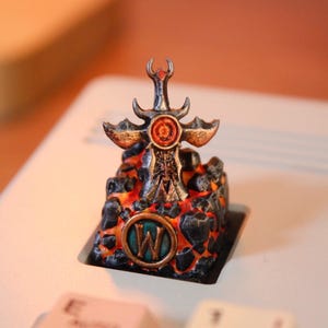 Puede incluir: Una tecla personalizada con un diseño de espada sobre una base de lava. La tecla tiene un acabado metálico con detalles en rojo, naranja y negro. La letra "W" es visible en la tecla.