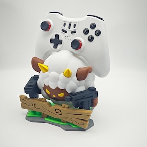 May include: A white and brown sheep figurine holding two black guns, standing on a wooden platform with green grass. The sheep is wearing a white and brown hat and has a black face. A white video game controller is mounted on the sheep's back.