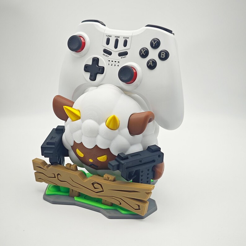 May include: A white and brown sheep figurine holding two black guns, standing on a wooden platform with green grass. The sheep is wearing a white and brown hat and has a black face. A white video game controller is mounted on the sheep's back.
