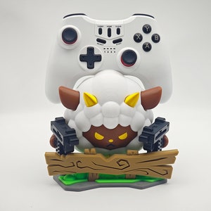May include: A white video game controller sits on top of a white sheep figurine holding two black guns. The sheep is standing on a brown wooden fence with green grass and a gray base.