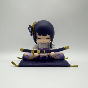 May include: A small purple and white figurine of a person sitting cross-legged on a purple cushion, holding a black sword. The figurine has black hair, a white face, and closed eyes. The figurine is wearing a white and purple outfit with gold accents.