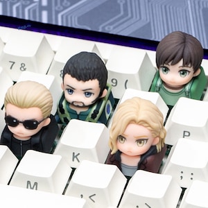 May include: A set of five custom keycaps featuring anime-style character designs. The keycaps are placed on a white keyboard, with the characters having detailed facial features and colorful outfits. The keycaps are a unique accessory for mechanical keyboards.