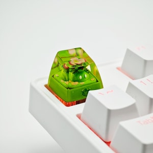 May include: A green and white resin keycap featuring a floral design and a small frog figurine. The keycap is placed on a white keyboard.
