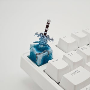 May include: A blue and white keycap with a silver sword sticking out of it. The keycap is on a white keyboard.