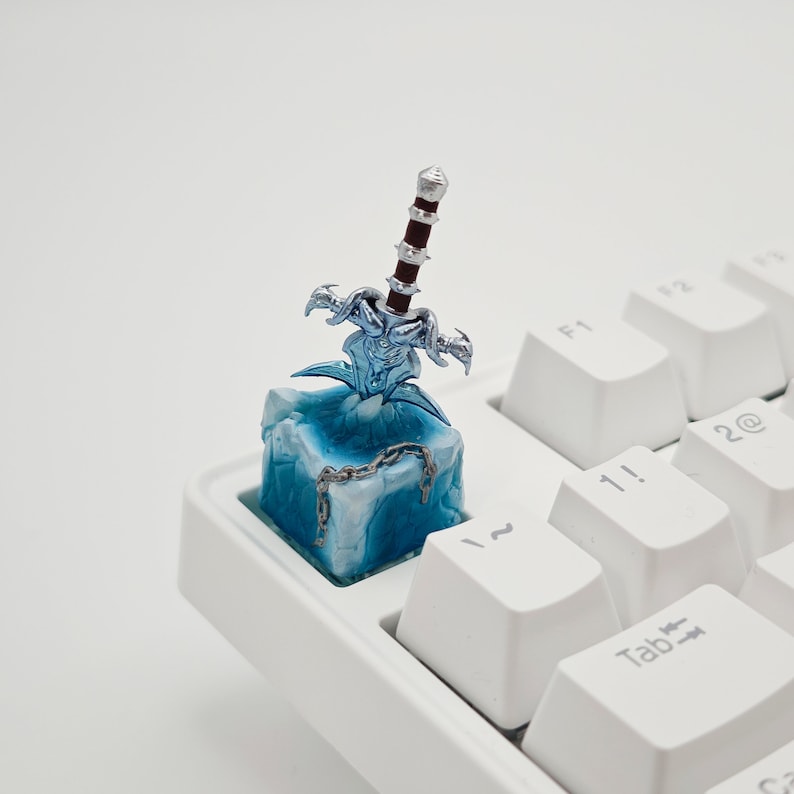 May include: A blue and white keycap with a silver sword sticking out of it. The keycap is on a white keyboard.