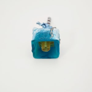 May include: A blue and white keycap with a chain and a yellow stem. The keycap is designed to look like a block of ice.