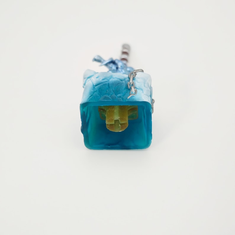 May include: A blue and white keycap with a chain and a yellow stem. The keycap is designed to look like a block of ice.