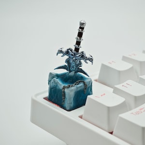 May include: A blue and silver keycap featuring a miniature sword with a dragon design. The keycap is on a white keyboard.