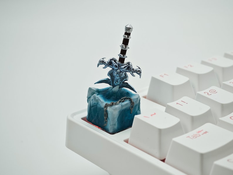 May include: A blue and silver keycap featuring a miniature sword with a dragon design. The keycap is on a white keyboard.