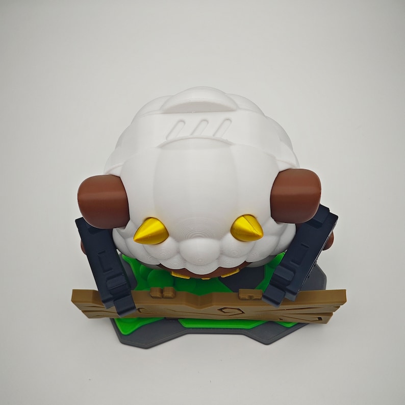 May include: A white sheep figurine with brown ears and gold horns, standing on a green and brown fence. The sheep is wearing a green shirt and has a black and white pattern on its body.
