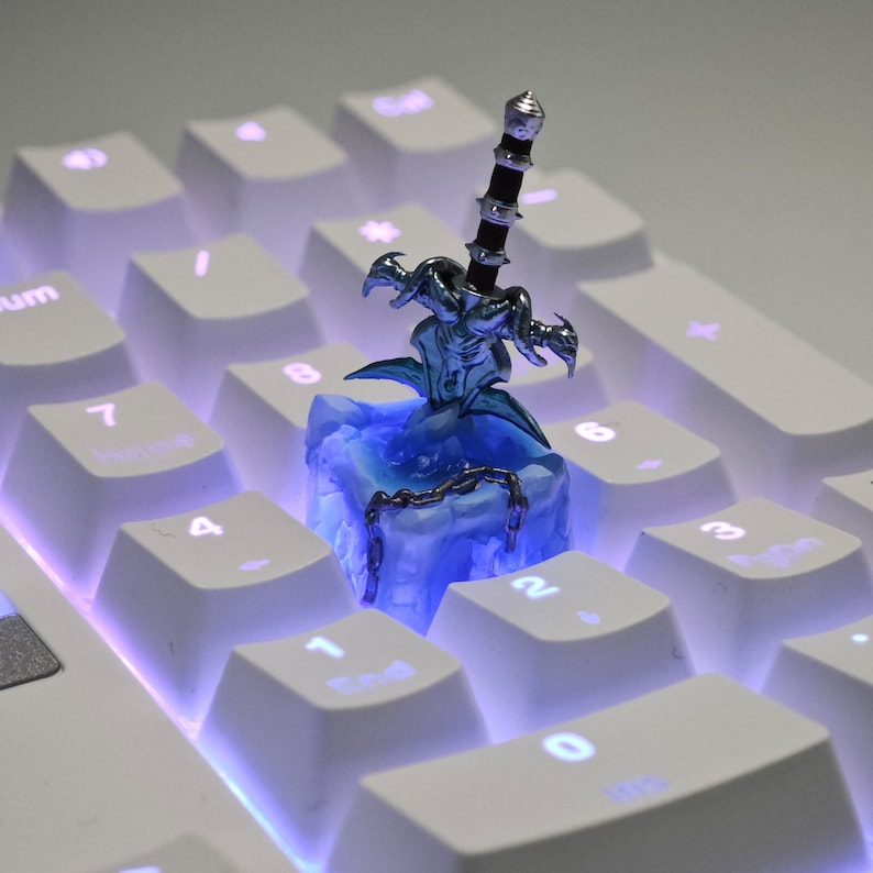 May include: A silver and blue miniature sword figurine on a blue and white ice-themed keycap. The keycap is on a white keyboard with blue backlighting.