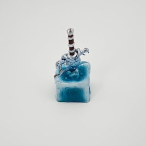 May include: A silver and red sword with a dragon design is frozen in a blue ice cube. The cube is made of resin and has a textured surface.