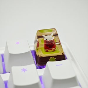Puede incluir: Un teclado blanco con una tecla que presenta un pequeño conejo blanco con un atuendo rojo y amarillo dentro de una tecla de resina transparente. La tecla está sentada en un teclado blanco con iluminación morada.