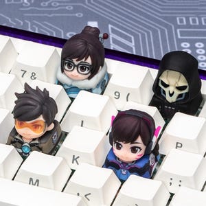 May include: Four custom artisan keycaps shaped like miniature character figures placed on a white mechanical keyboard. The figures include a character with dark hair and glasses, a hooded skull figure, a character with orange goggles, and a character with pink headset accents.