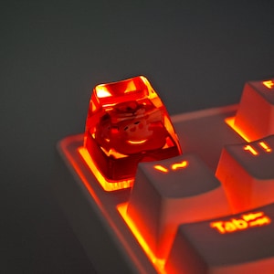 May include: A red transparent keycap with a design inside, lit up from below, on a white keyboard with the keys "/" and "Tab" visible.