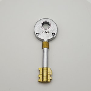 Könnte beinhalten: Ein silberner Schlüssel mit einem runden Kopf und einem goldfarbenen Schlüsselbart. Der Kopf des Schlüssels hat ein kleines Loch in der Mitte und den Text "K-24A" aufgedruckt.