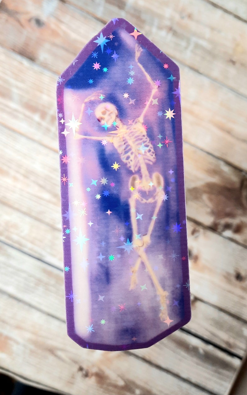 May include: A purple crystal-shaped sticker with a holographic finish features a skeleton dancing inside. The background is filled with iridescent stars.