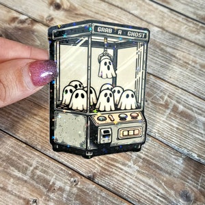 May include: A vintage-style sticker of a ghost claw machine with the text "Grab a Ghost" on the top. The machine is filled with cartoon ghosts and has a claw reaching down to grab one.