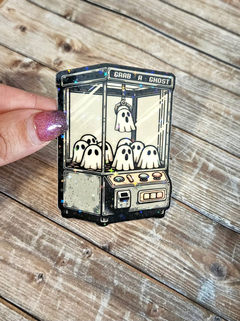 May include: A vintage-style sticker of a ghost claw machine with the text "Grab a Ghost" on the top. The machine is filled with cartoon ghosts and has a claw reaching down to grab one.