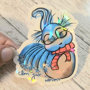 May include: A holographic sticker featuring a whimsical cartoon worm with a blue body, a coral scarf, and closed eyes. The sticker has the text "I'm just a worm" in a handwritten style.