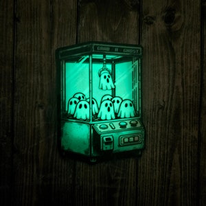 May include: A green glow-in-the-dark sticker of a vintage claw machine with the text "Grab a Ghost" on the front. The claw machine is filled with cartoon ghosts.