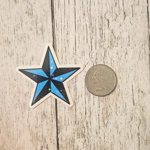 May include: A star-shaped sticker with a black and blue design. The sticker has a white border and is next to a quarter coin for size comparison. The star has a blue center with black points and small, colorful accents.