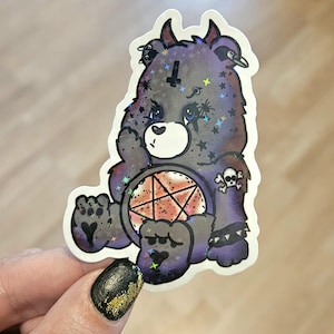 May include: A holographic sticker featuring a dark purple bear with a pentagram, stars, and skull accents. The bear has a cross symbol on its forehead and is holding a pentagram. The sticker has a white border.