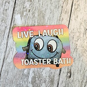 May include: A holographic sticker featuring a cartoon toaster with large eyes and a smiling mouth. The background is a rainbow gradient with a star pattern. The text "LIVE, LAUGH TOASTER BATH" is printed in white capital letters above and below.