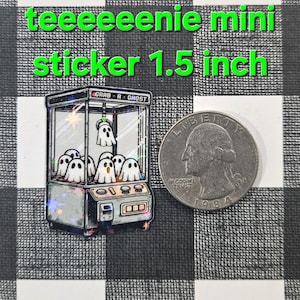 May include: A holographic sticker of a ghost claw machine with the text "Grab-A-Ghost" on the top. The sticker is 1.5 inches in size and is next to a US quarter for size comparison.