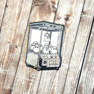 May include: A black and white illustration of a claw machine with the text "Grab-A-Ghost" on the top. The claw machine is filled with ghosts.