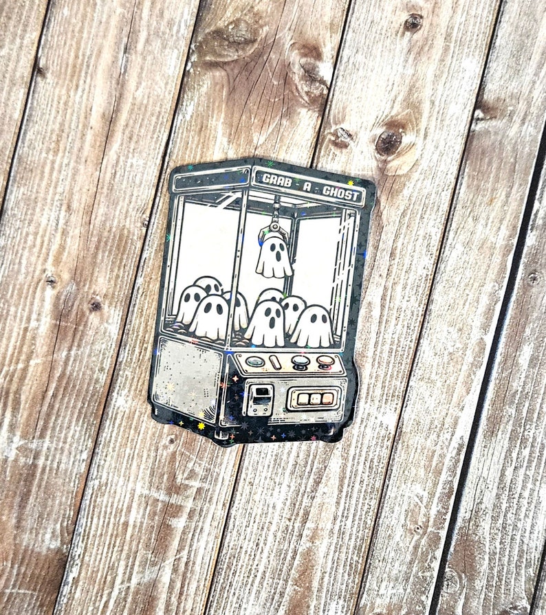 May include: A black and white illustration of a claw machine with the text "Grab-A-Ghost" on the top. The claw machine is filled with ghosts.