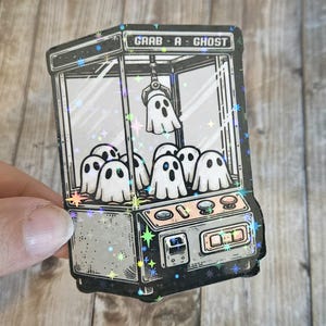May include: A sticker of a vintage-style claw machine with the text "Grab-A-Ghost" on the front. The machine is filled with ghosts, and there are stars and sparkles around it.