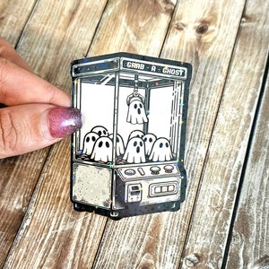 May include: A black and white sticker of a ghost claw machine with the text "Grab a Ghost". The machine is filled with ghosts and has a starry background.