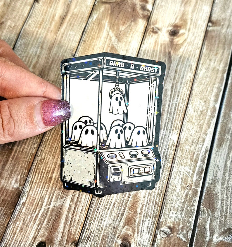 May include: A black and white sticker of a ghost claw machine with the text "Grab a Ghost". The machine is filled with ghosts and has a starry background.