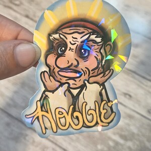 May include: Holographic sticker featuring a cartoon character with a sun-like halo. The character has a detailed face with wrinkles and a stern expression. The word "Hobble" is written in a stylized font.