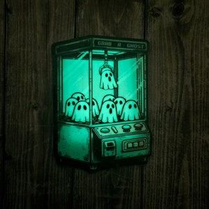 May include: A green glow-in-the-dark sticker of a ghost claw machine with the text "Grab a Ghost" on the top. The claw is holding a ghost and there are multiple ghosts inside the machine.