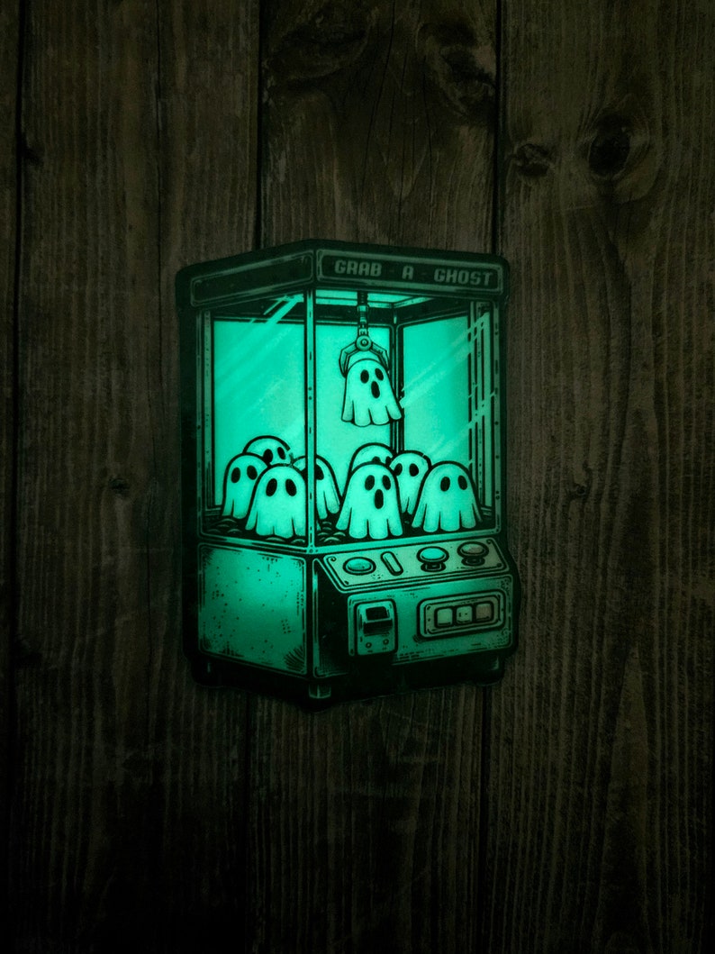 May include: A green glow-in-the-dark sticker of a ghost claw machine with the text "Grab a Ghost" on the top. The claw is holding a ghost and there are multiple ghosts inside the machine.